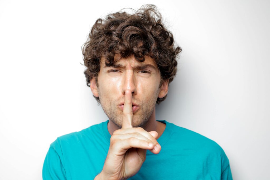 Adult man with curly hair in blue shirt making a silence gesture on white background.